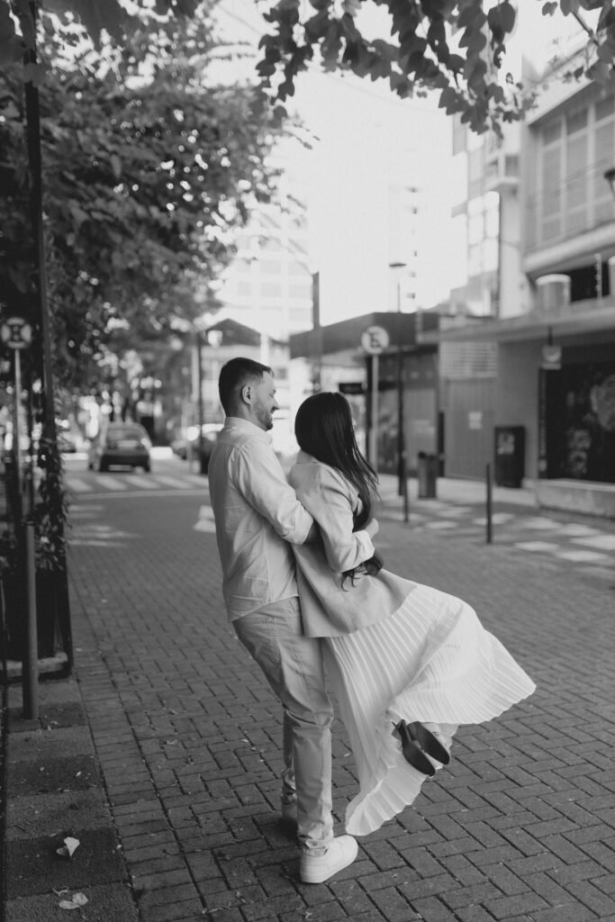 A black and white photo of a couple embracing joyfully on a city street.